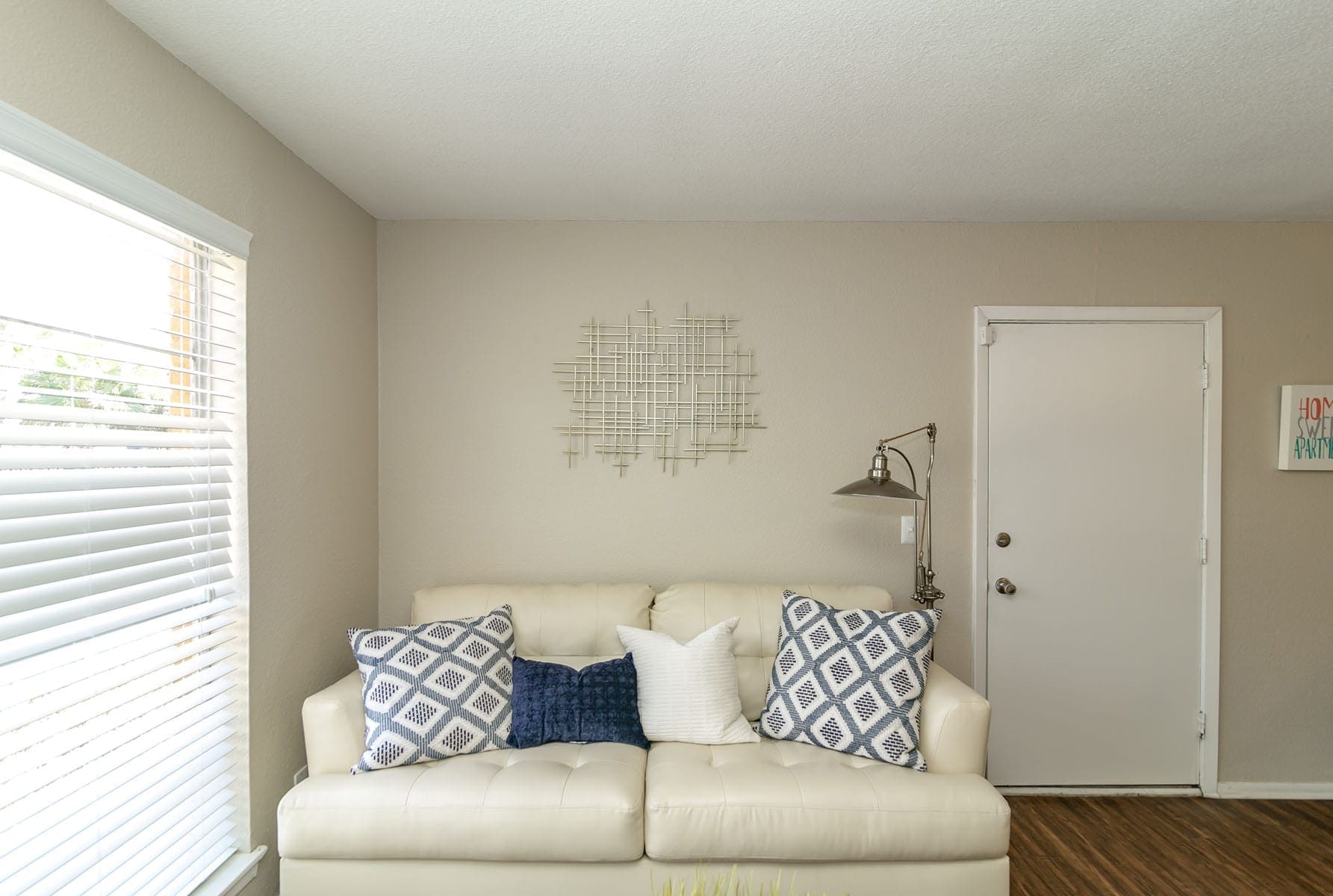 Whit couch with blue, grey and white pillows. Windoe to the left and a door to the right. A white wall in the background.